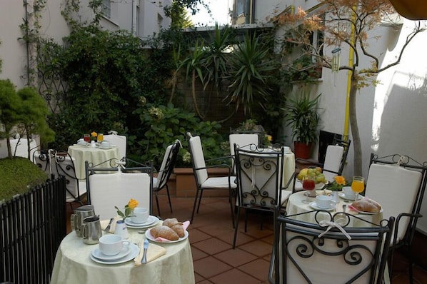 Une terrasse extérieure avec des tables dressées pour le repas, entourée de verdure et bordée d’un bâtiment blanc.