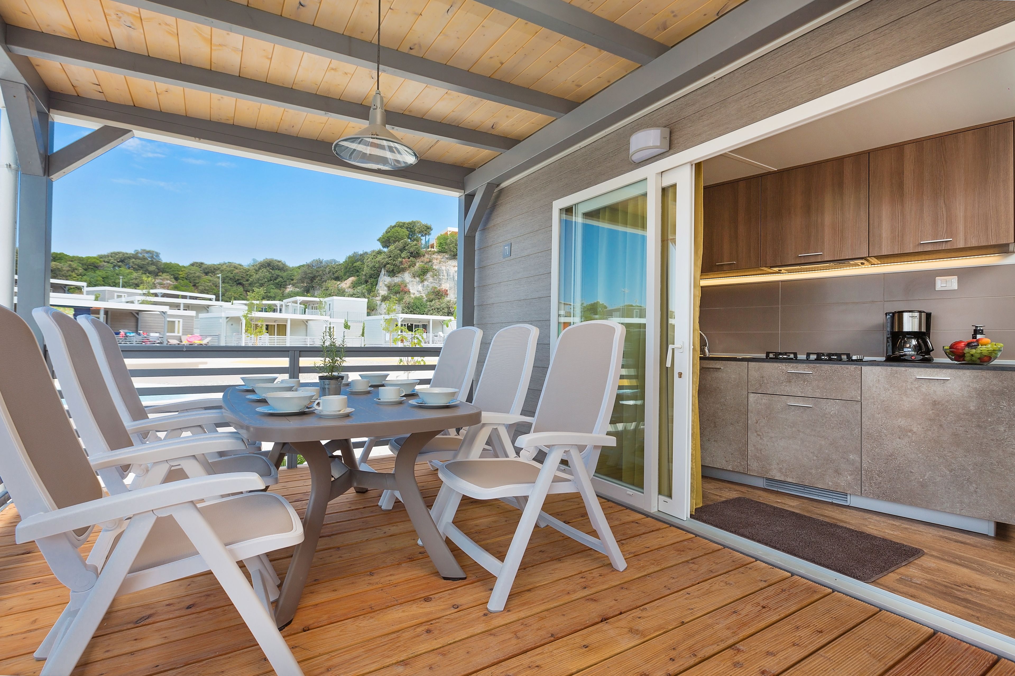 A covered outdoor dining area with a table and chairs, a kitchen with cabinets and appliances, and a view of the surrounding landscape.