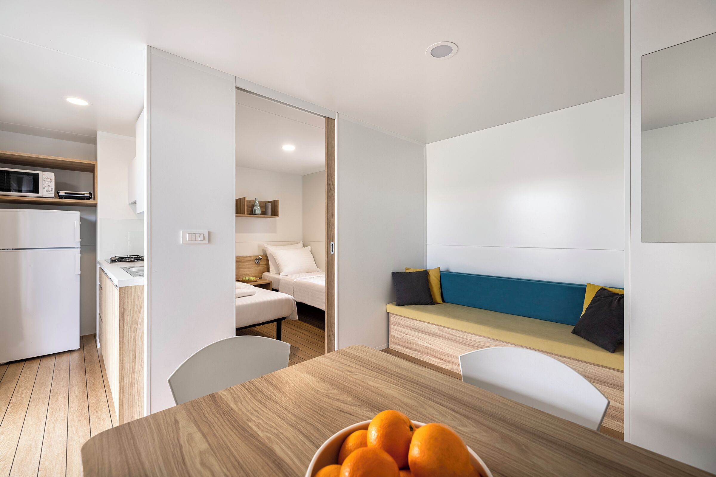 A compact living space with a kitchenette, a dining area with a bowl of oranges, and a seating area with a sofa and cushions.