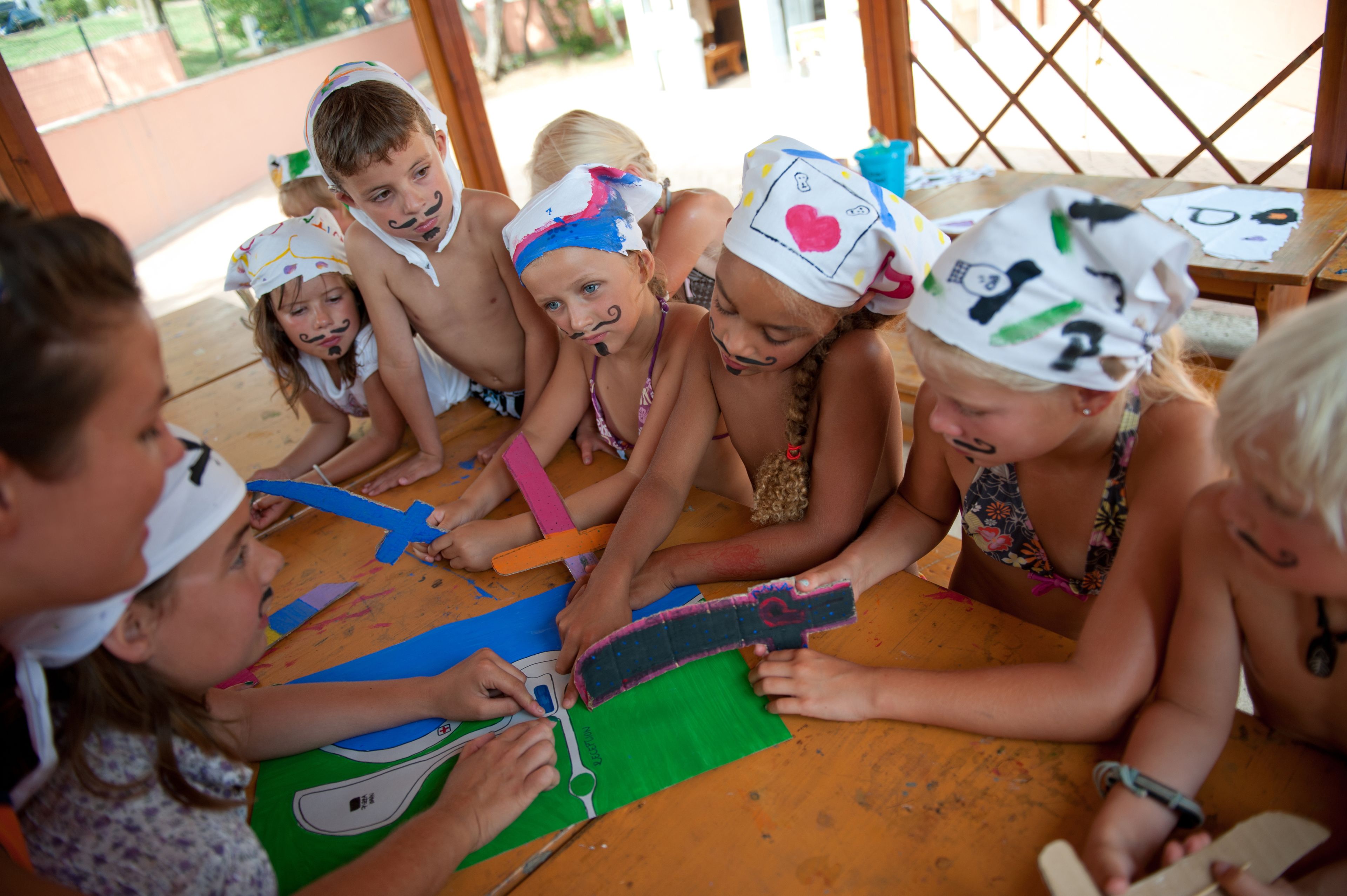 Eine Gruppe Kinder ist mit Bastelarbeiten an einem Tisch beschäftigt, auf dem verschiedene farbige Papiere und Werkzeuge liegen.