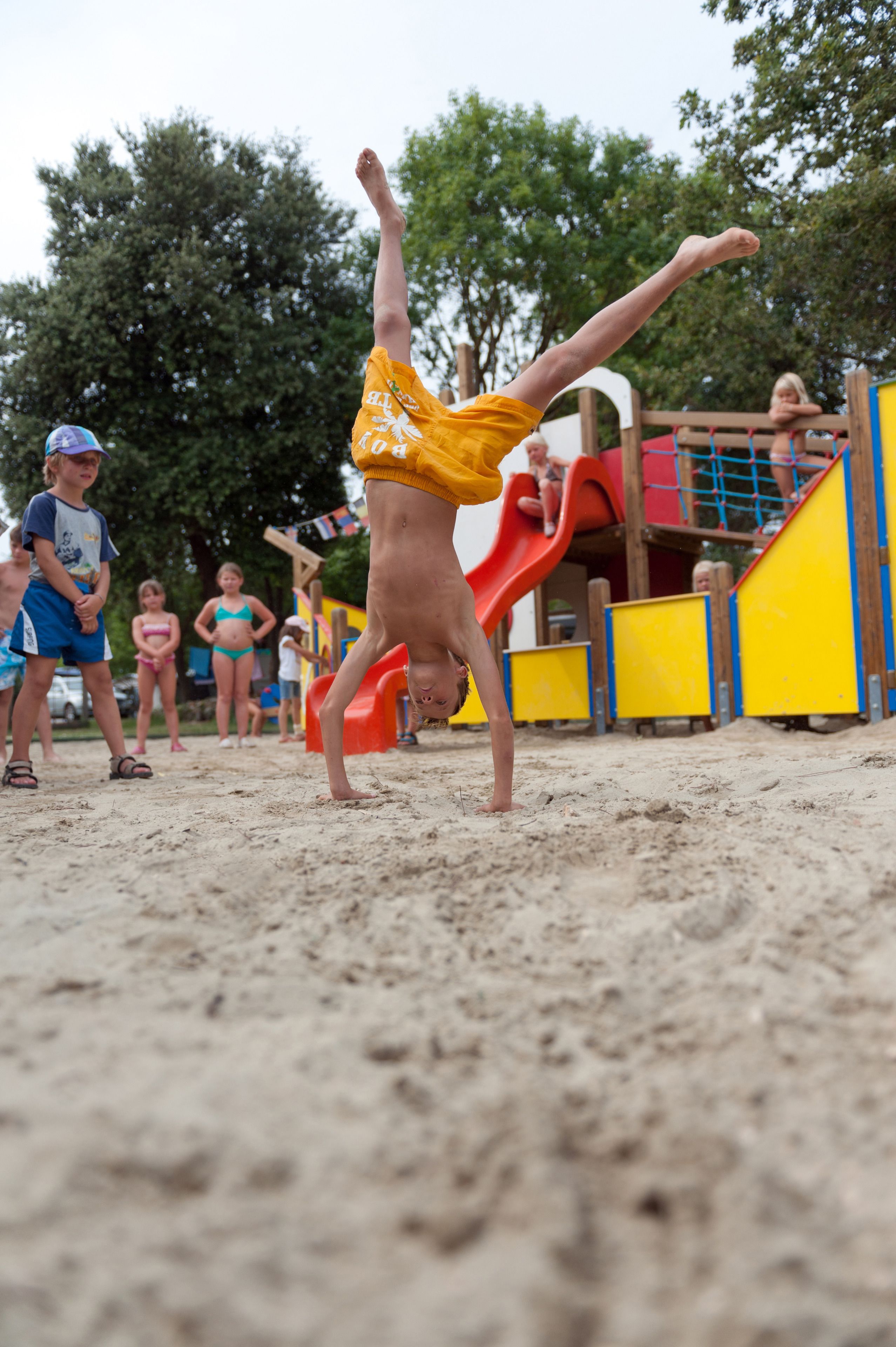 A person performing a handstand on a sandy surface with playground equipment in the background.