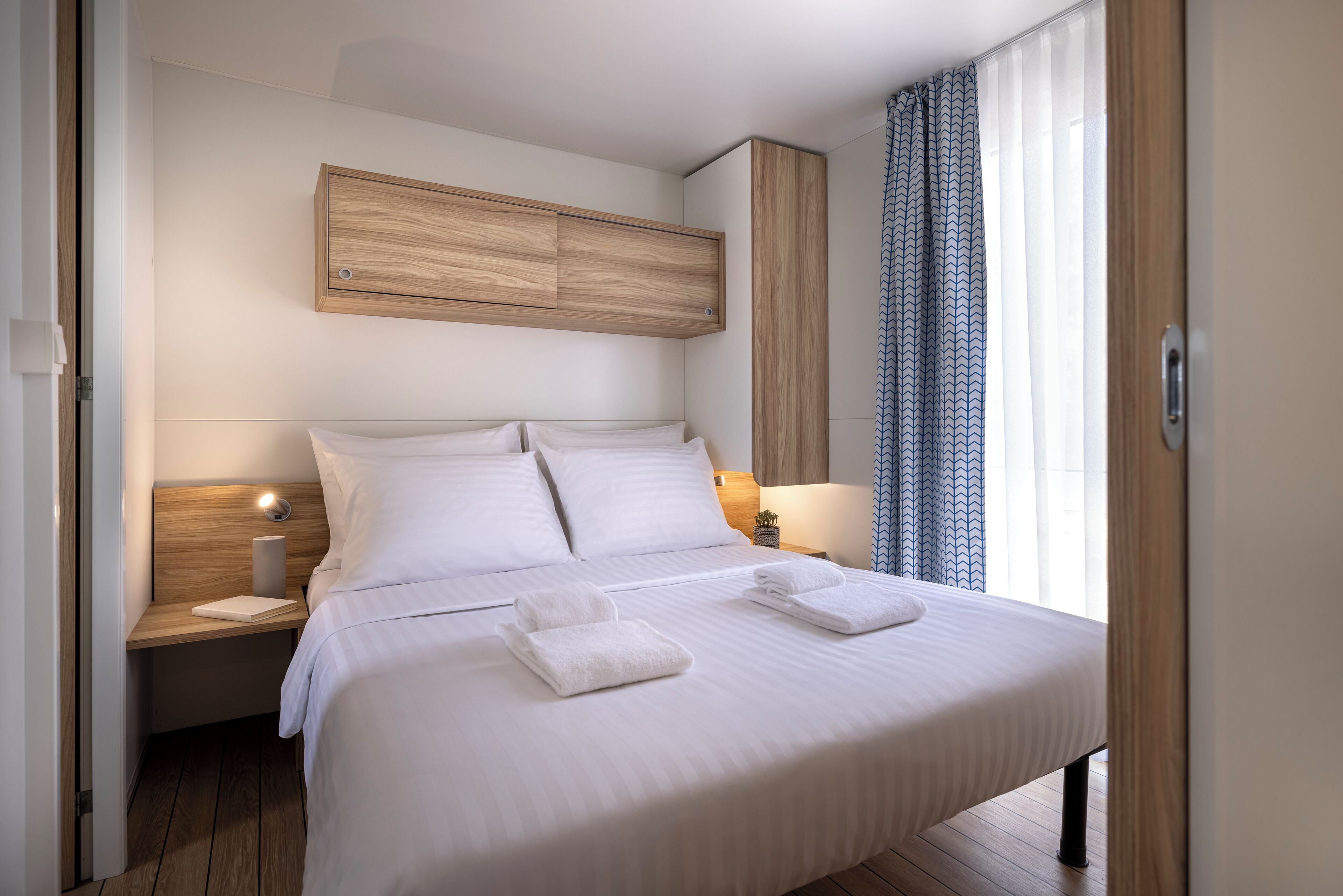 A neatly made bed with white linens and two folded towels, a wooden headboard, a bedside table with a lamp, and a window with patterned curtains.