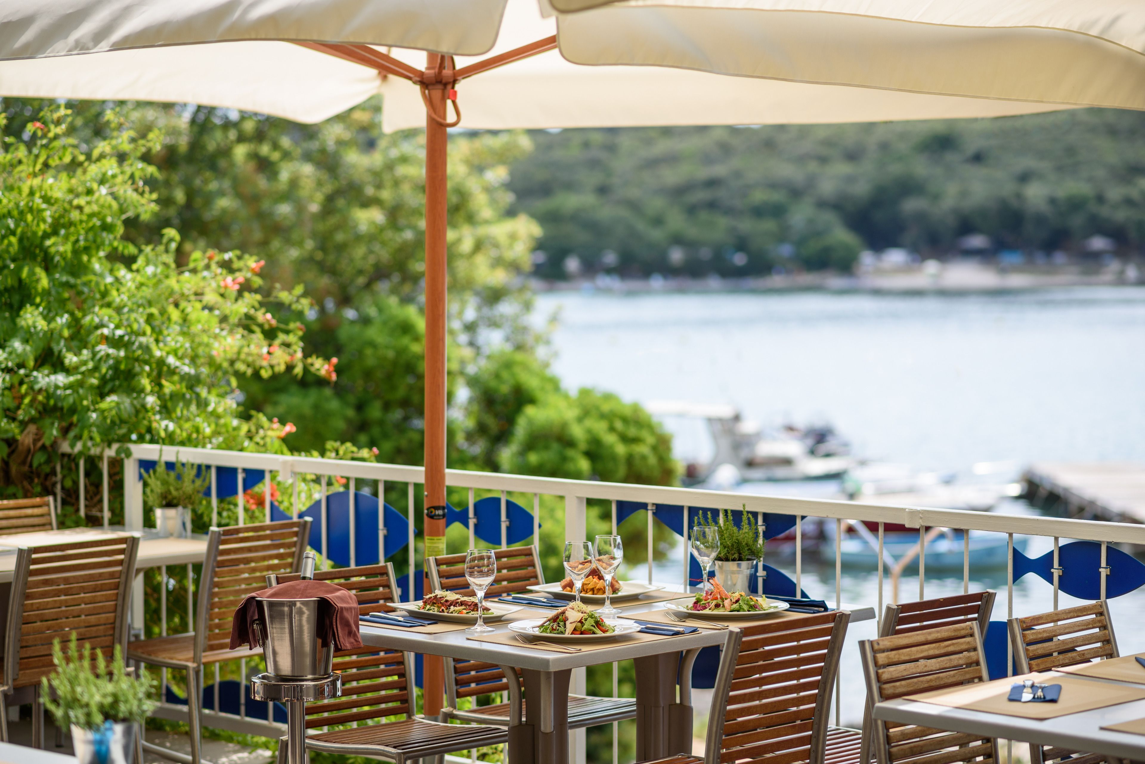 Un espace extérieur aménagé pour les repas, avec des tables et des chaises, une vue sur l’eau et une rambarde ornée de motifs bleus et blancs.