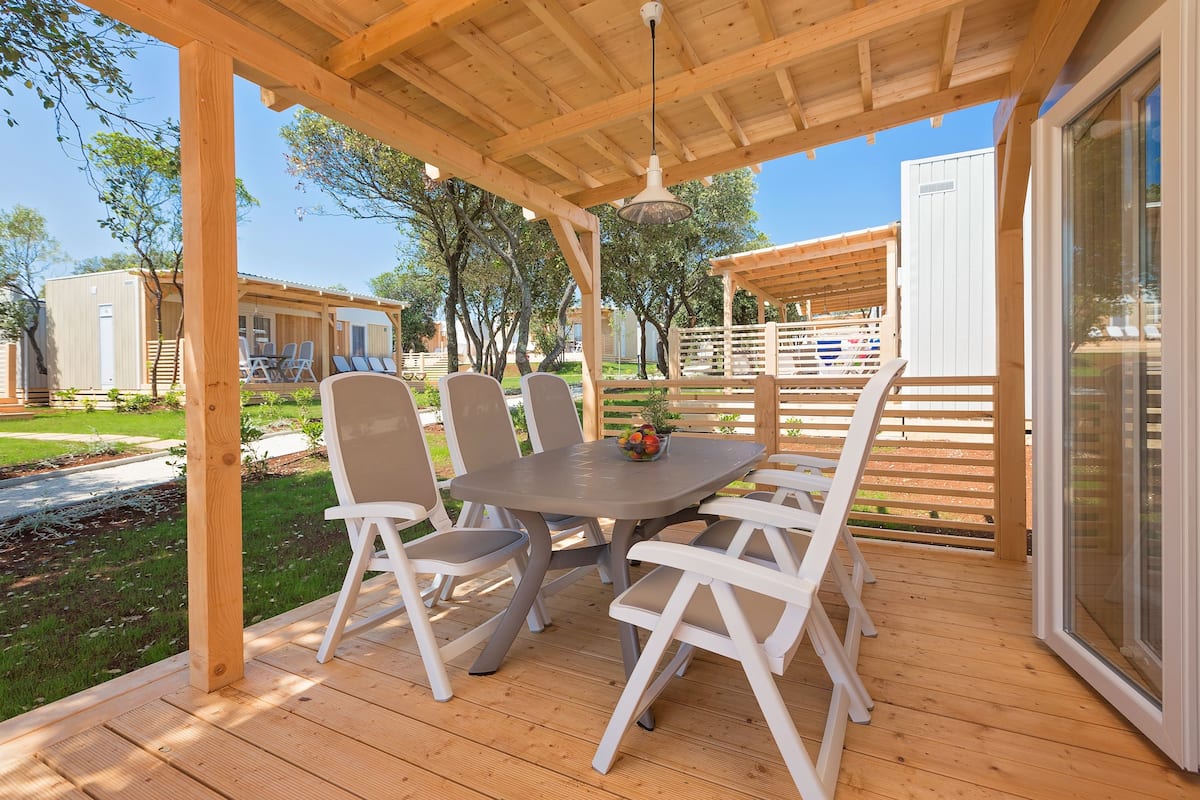 A covered outdoor patio with a table and chairs, surrounded by wooden deck and a view of mobile homes.