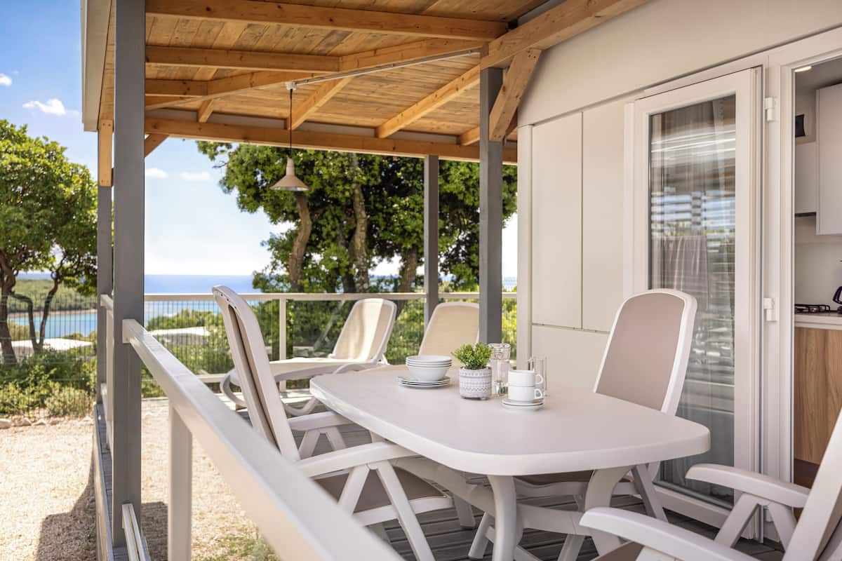 A covered outdoor dining area with white furniture, a view of the sea, and a kitchen area with a coffee maker.
