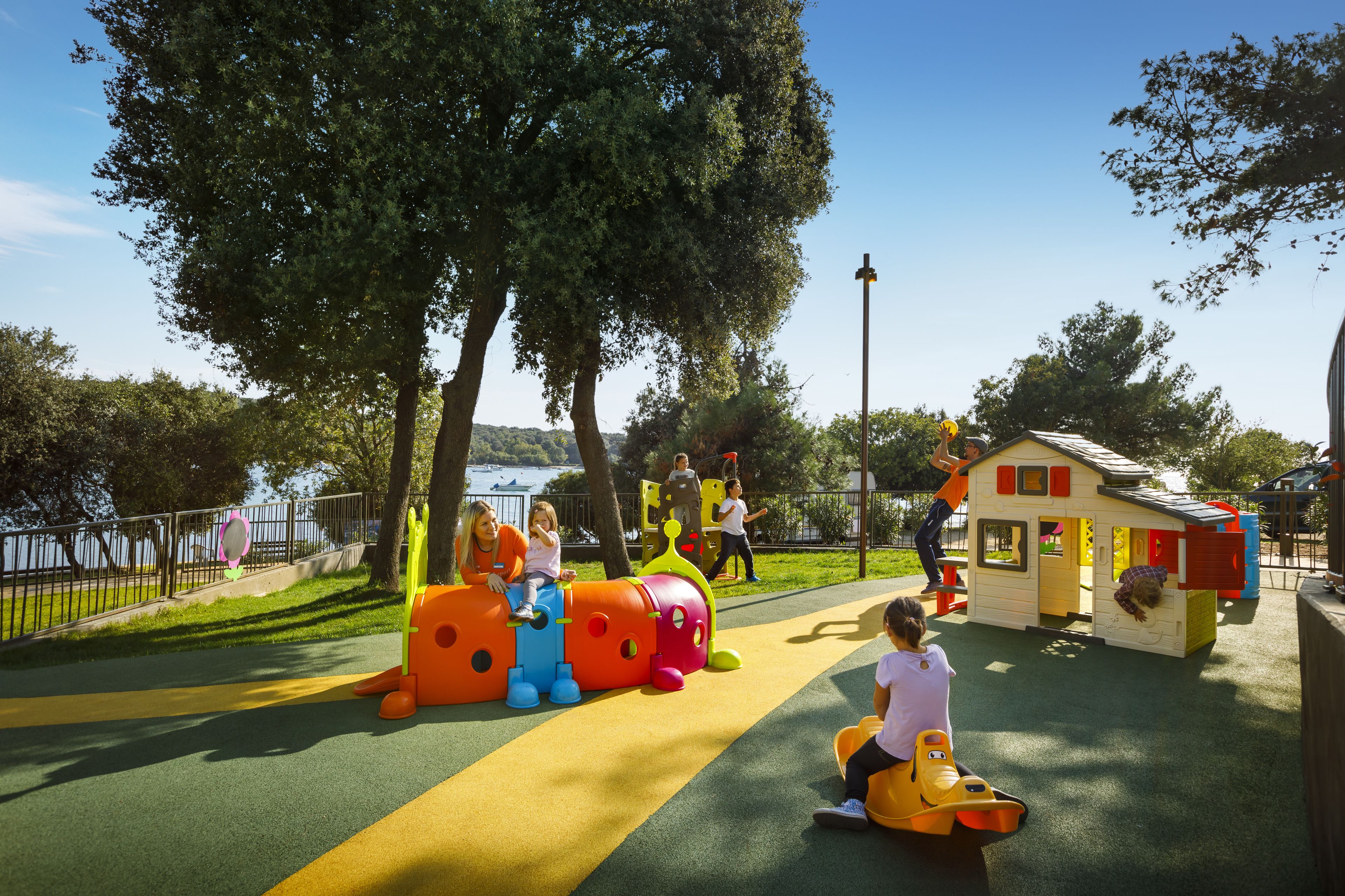 A playground with a colorful slide and play structures, surrounded by trees and a body of water in the background.