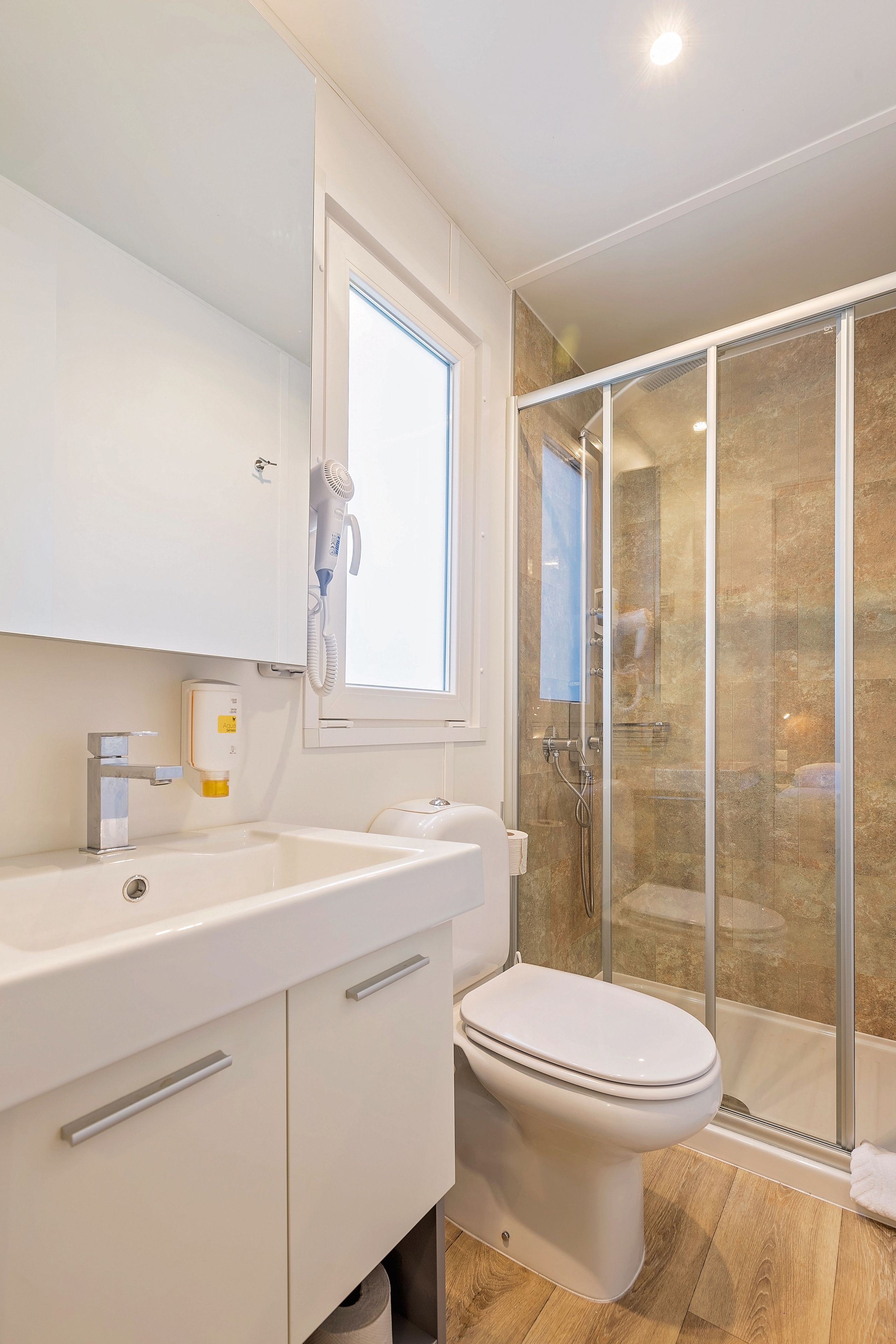 A modern bathroom with a white vanity, a toilet, a glass-enclosed shower, and a window providing natural light.