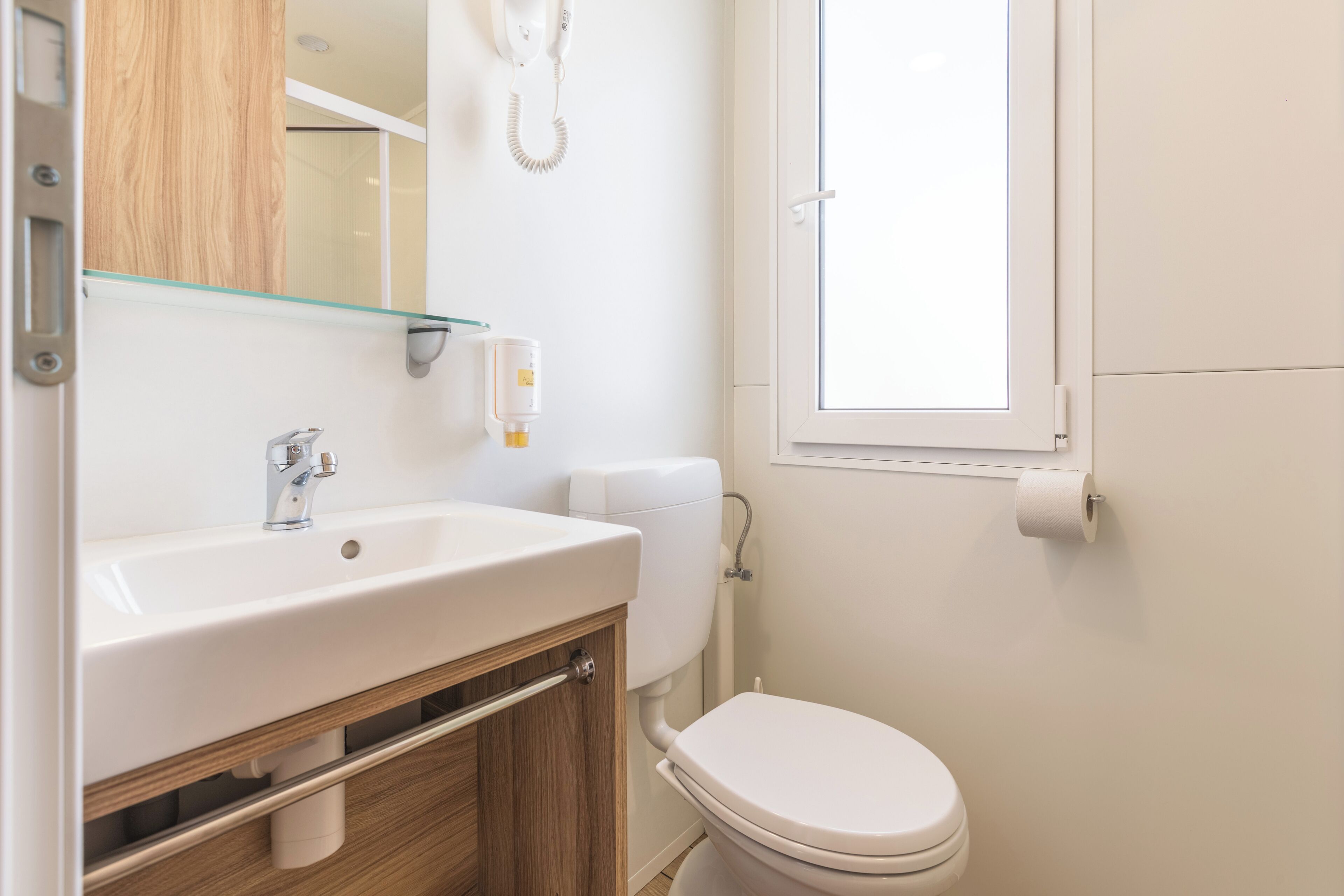 Un baño con lavamanos blanco, mueble de madera, inodoro y ventana.