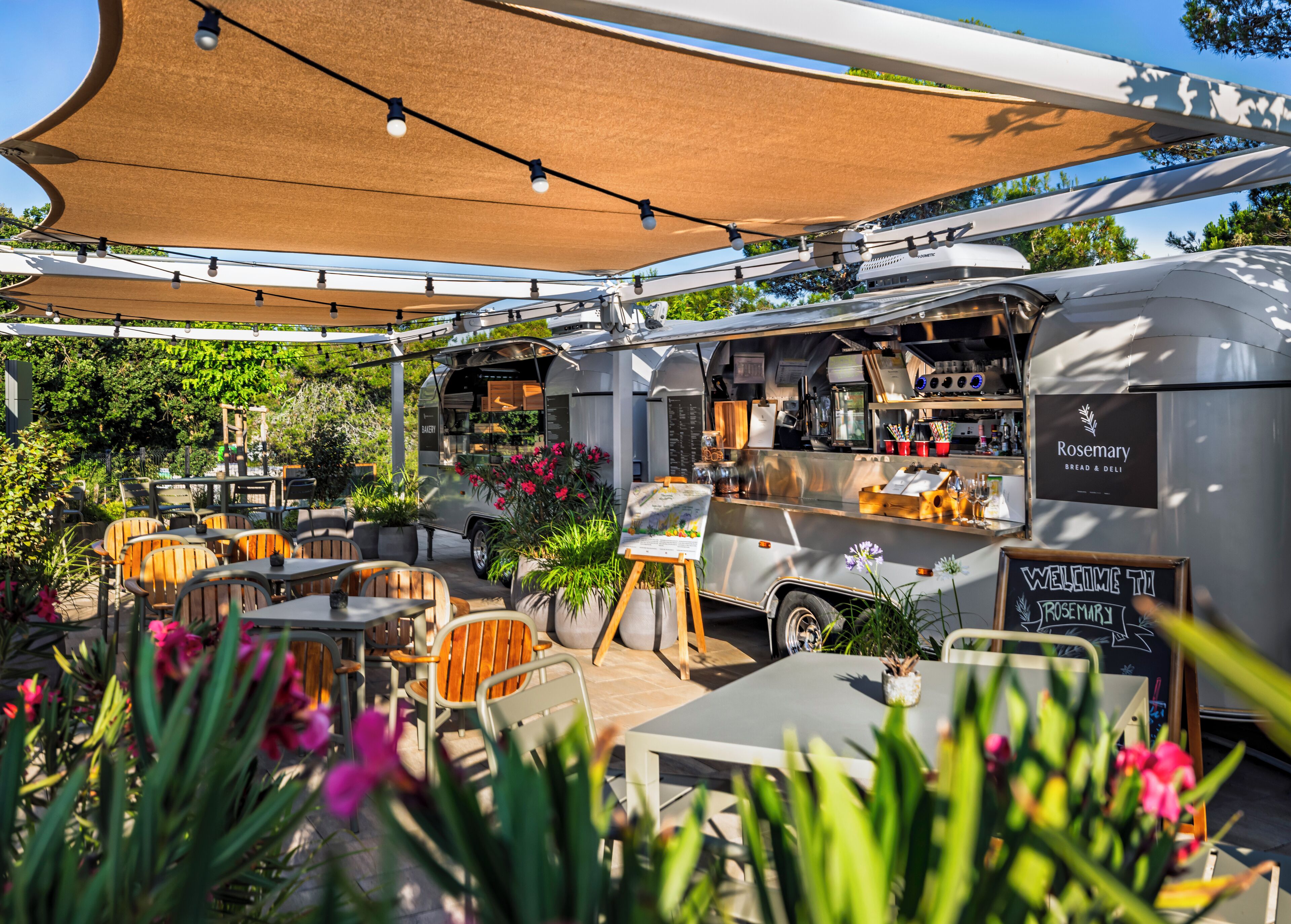A food truck with a sign reading "Rosemary Bread & Deli" parked in an outdoor seating area with tables and chairs, surrounded by greenery.