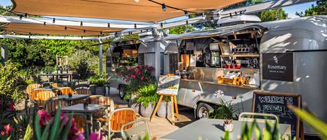 A food truck with a sign reading "Rosemary Bread & Deli" parked in an outdoor seating area with tables and chairs, surrounded by greenery.