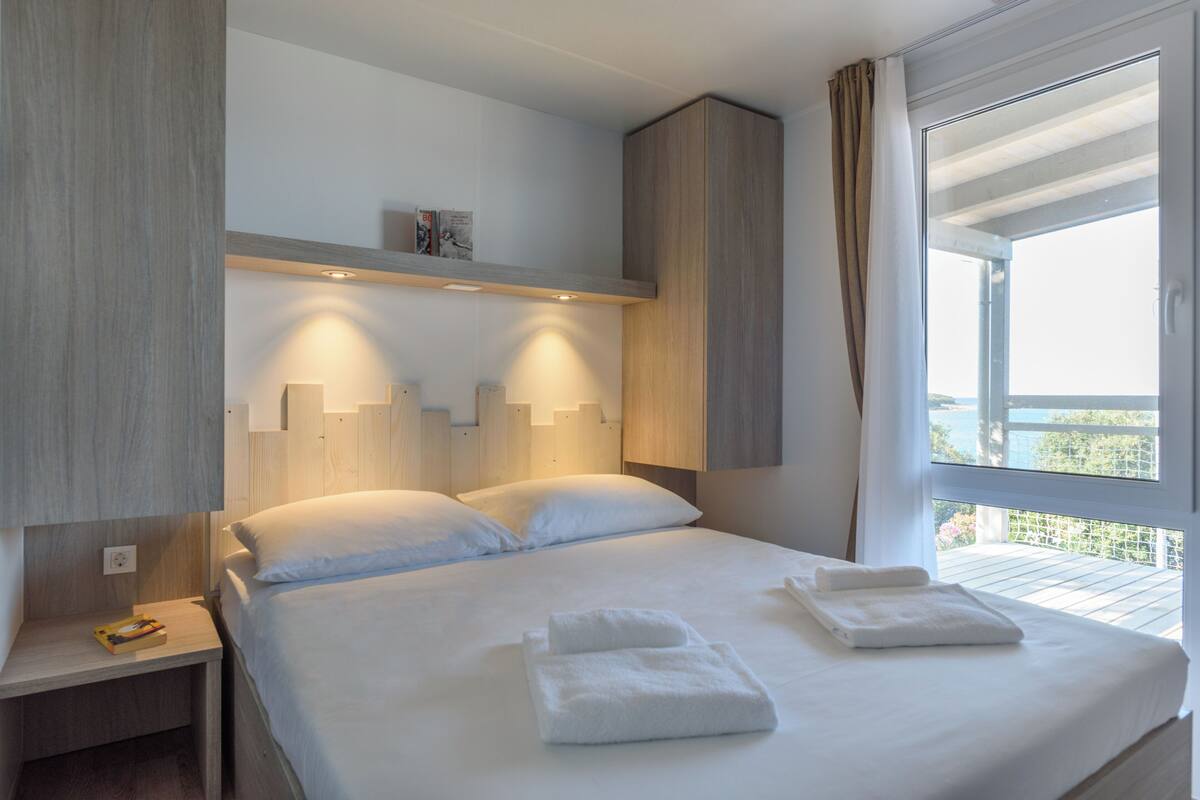 A hotel room with a large bed, two bedside tables, a nightstand, a built-in shelf with books, and a window with curtains.