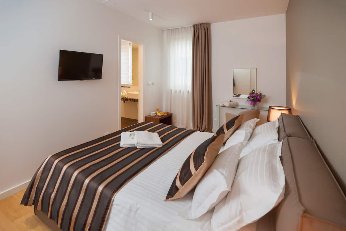 A hotel room with a bed, a TV mounted on the wall, a bedside table with a lamp, and a bathroom visible through an open door.