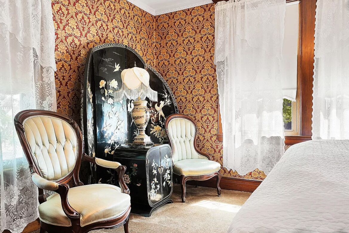 A corner room with two upholstered chairs, a large mirror with floral designs, and a patterned wallpaper.