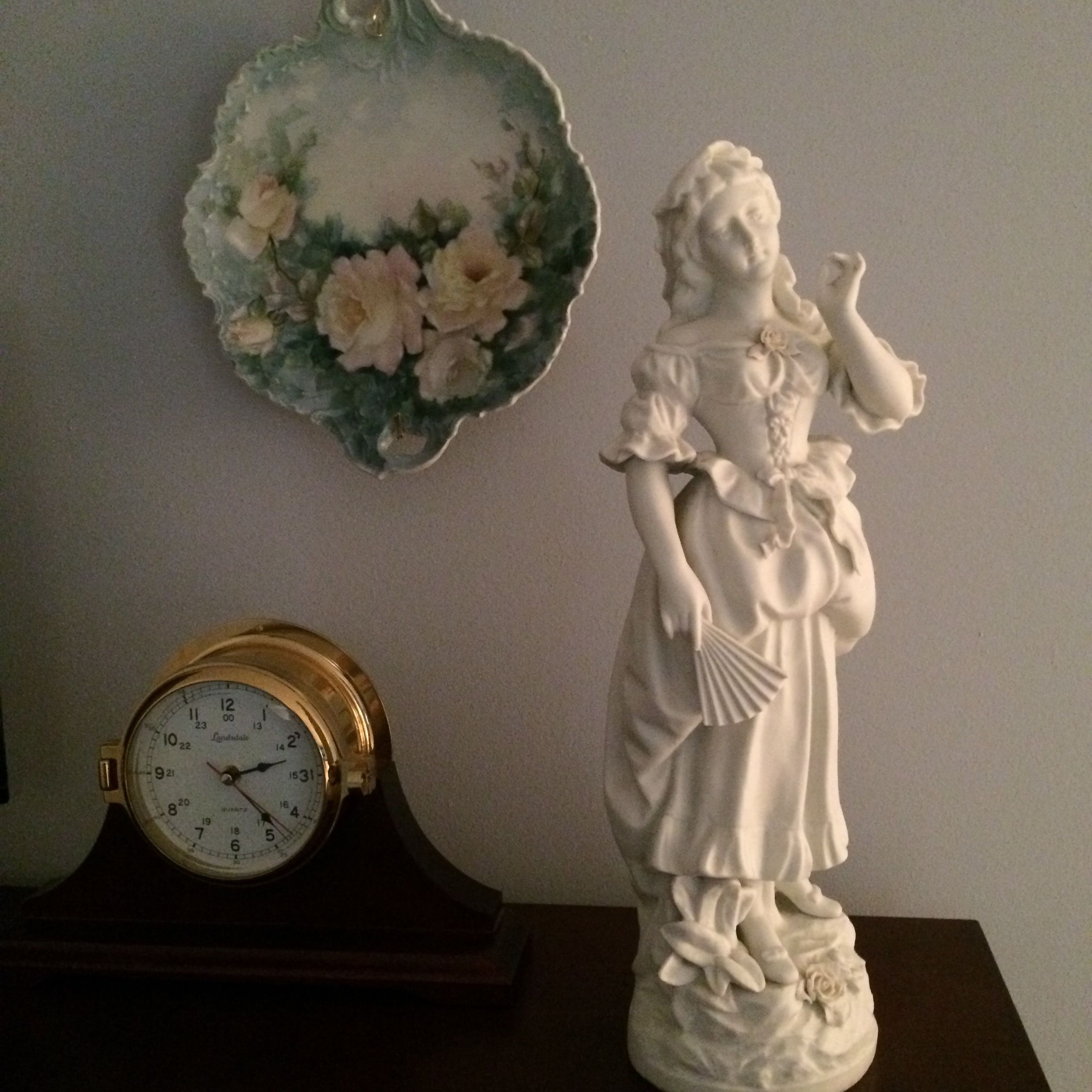 Un reloj de pared, un plato decorativo con motivos florales y una figurita blanca.