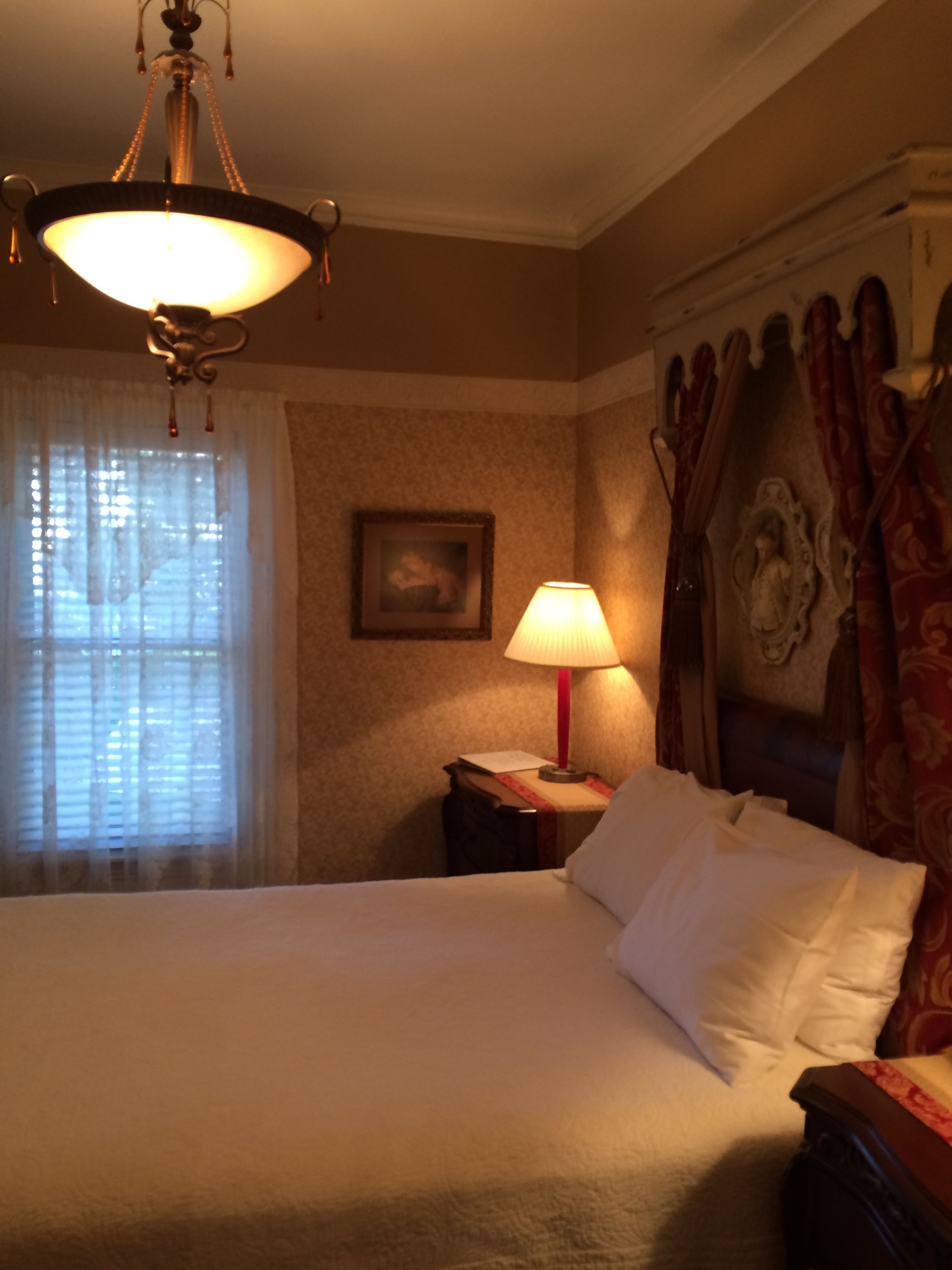 A hotel room with a bed, a nightstand, a lamp, a chandelier, curtains, and a framed picture on the wall.