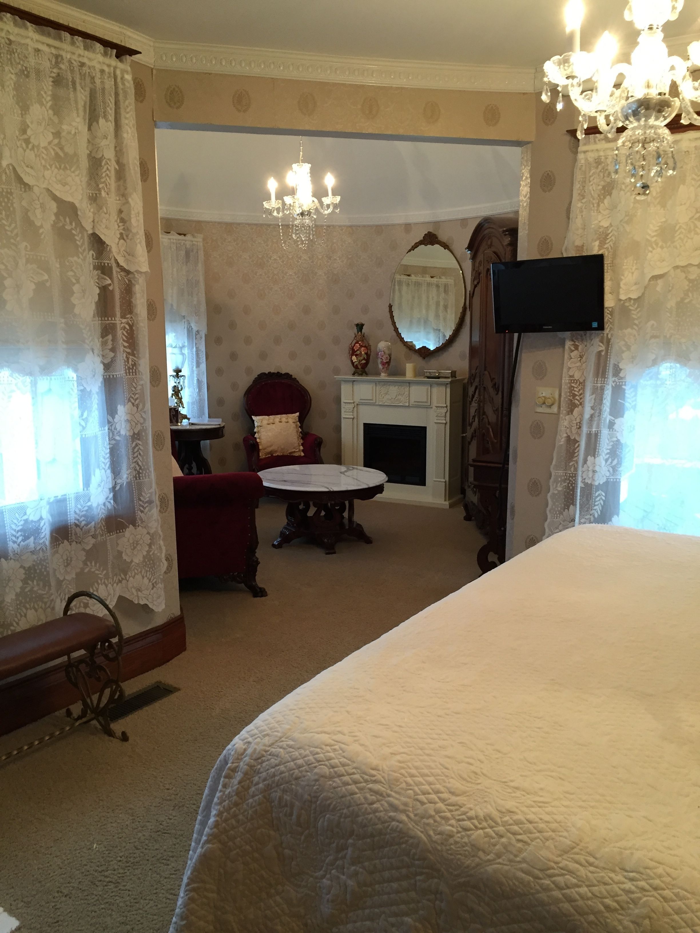 A bedroom with a large bed, a round table, a red armchair, a fireplace, and a television.