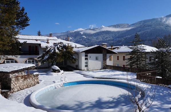 Piscina al aire libre cubierta de nieve, rodeada de edificios con montañas al fondo.