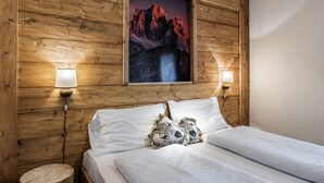Une chambre à coucher avec une tête de lit en bois, un tableau encadré représentant une montagne, deux lampes de chevet et un lit recouvert de draps blancs et agrémenté de coussins décoratifs.