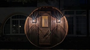 Une structure en forme de tonneau, faite de bois, avec une porte et deux fenêtres, éclairée par des lampes.