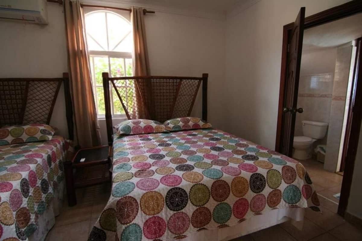 Une chambre avec deux lits simples, une tête de lit en bois, une fenêtre avec des rideaux et une porte donnant sur une salle de bain.