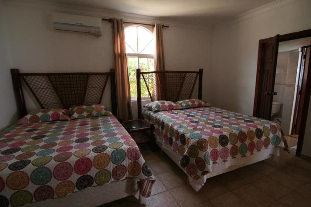 A bedroom with two beds, each with a colorful circular-patterned bedspread. There is a window with curtains, an air conditioning unit on the wall, and a door to the right.