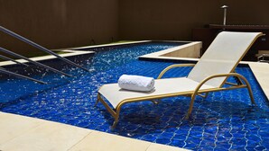 Una piscina con suelo de mosaico azul, una toalla blanca en una silla de descanso y una escalera metálica.