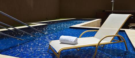 A swimming pool with a blue mosaic floor, a white towel on a lounge chair, and a metal ladder.