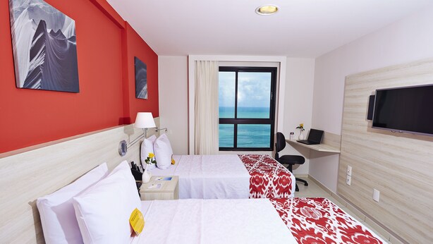 Habitación de hotel con dos camas, un escritorio, una silla, un televisor y un ventanal con vistas al mar.