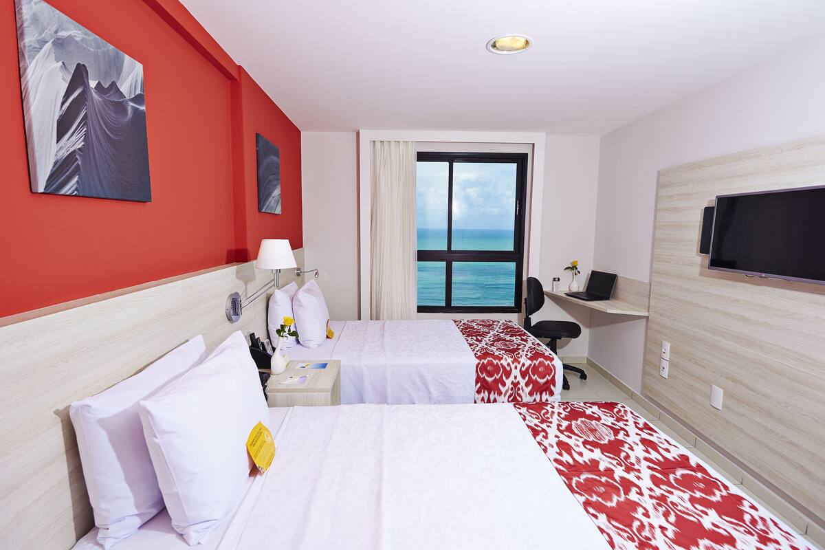 A hotel room with two beds, a desk, a chair, a TV, and a large window with a view of the sea.