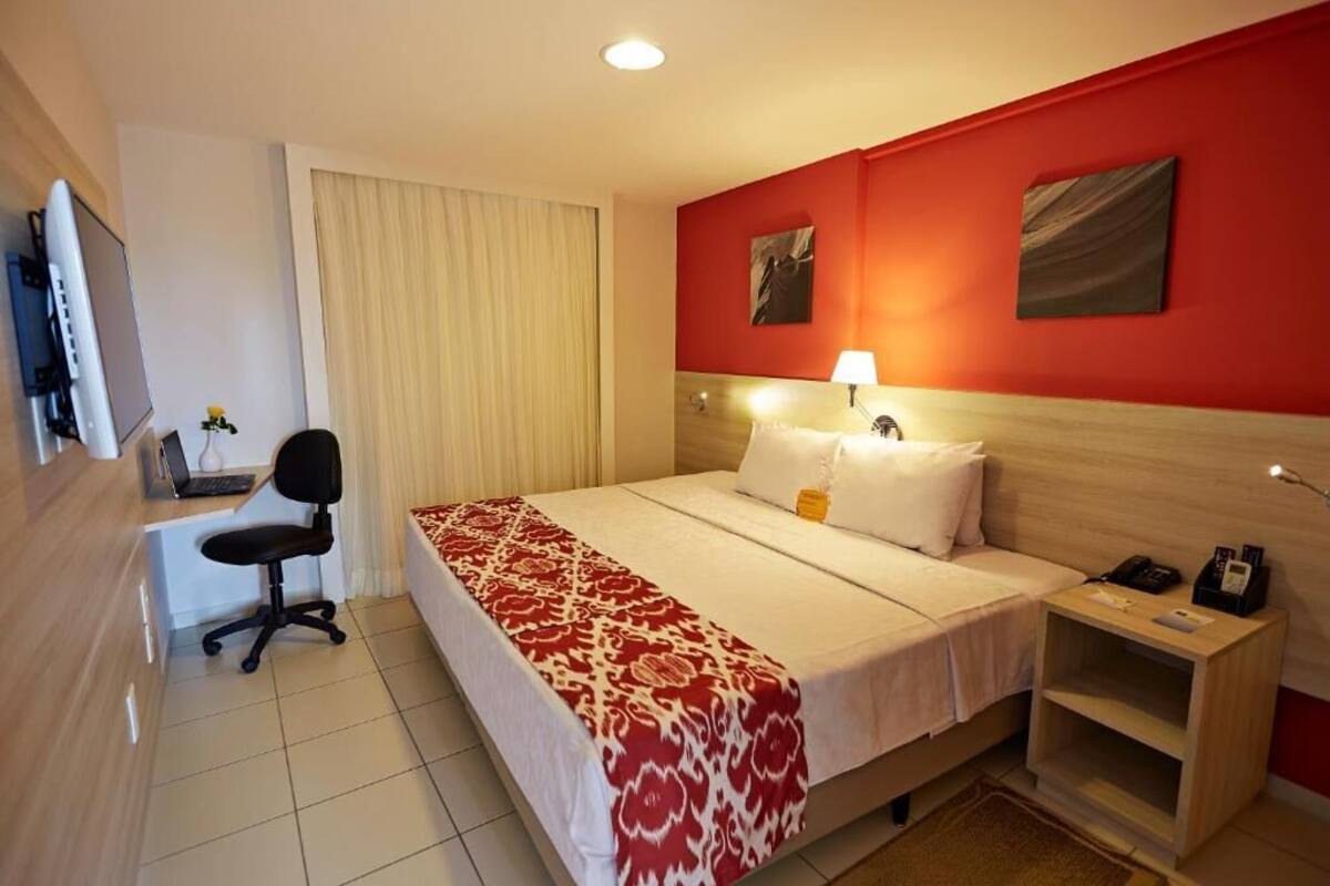 A hotel room with a bed, a desk with a chair, a bedside table, and a red accent wall.