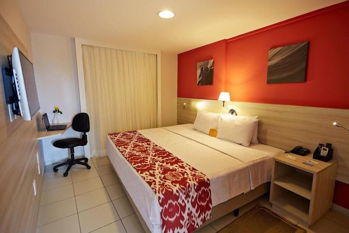 A hotel room with a bed, a desk with a chair, a bedside table, and a red accent wall.
