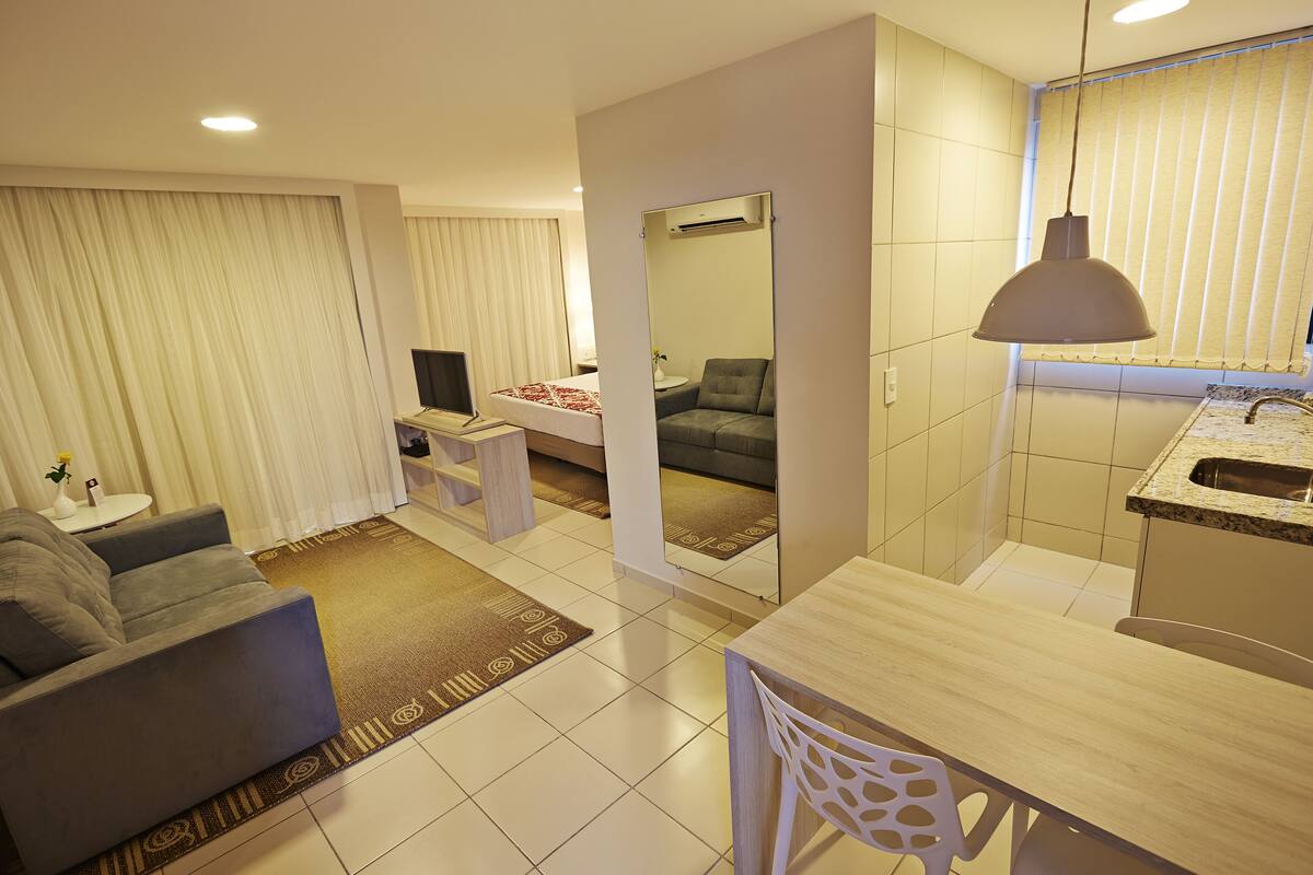 A modern living room with a grey sofa, a small white table, and a TV. There is a dining area with a wooden table and chairs. The room has a large mirror and a ceiling light.