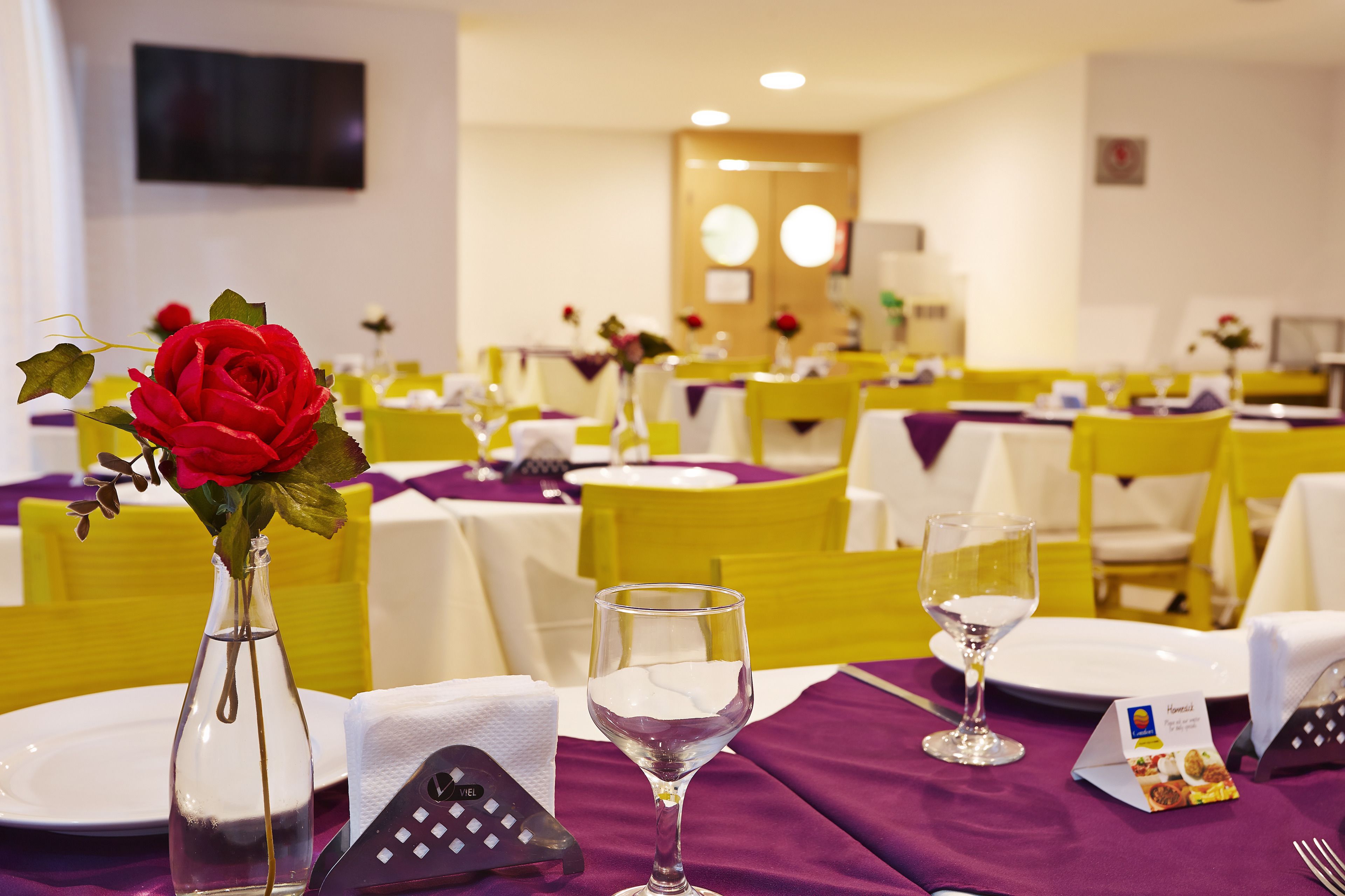 A dining area with yellow chairs, white tables, and purple tablecloths. There is a vase with a red rose and a wine glass on the table.