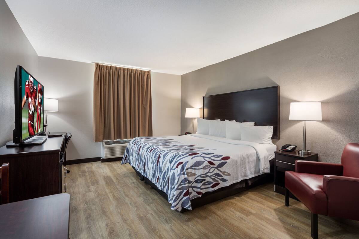 Une chambre d’hôtel avec un lit, des lampes de chevet, un bureau avec un ordinateur, une télévision et un fauteuil rouge.