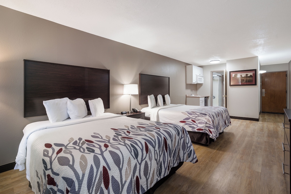 Habitación de hotel con dos camas, una mesita de noche con lámpara, un espejo y una puerta que da a otra habitación.