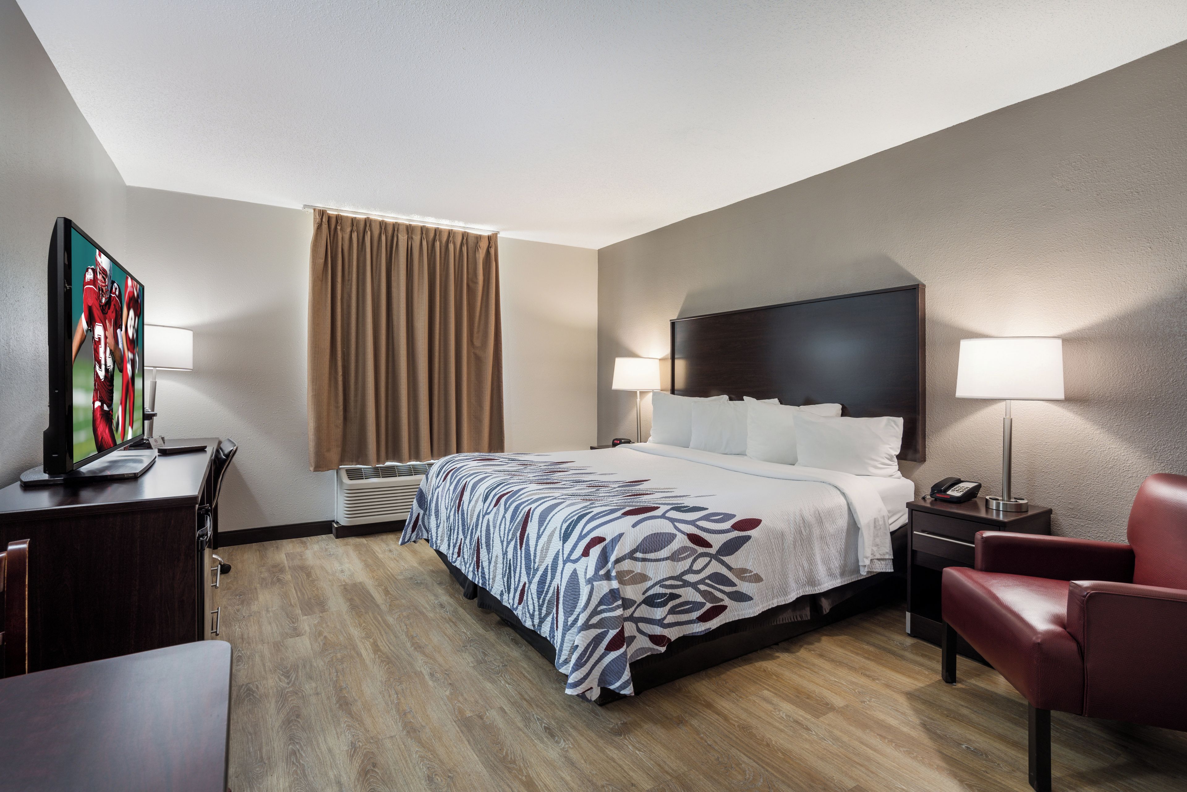 A hotel room with a bed, a desk with a computer, a chair, a TV, and a window with curtains.