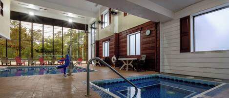 Una piscina interior con suelo de baldosas, un pequeño espacio de descanso con sillas y vistas a árboles a través de grandes ventanales.