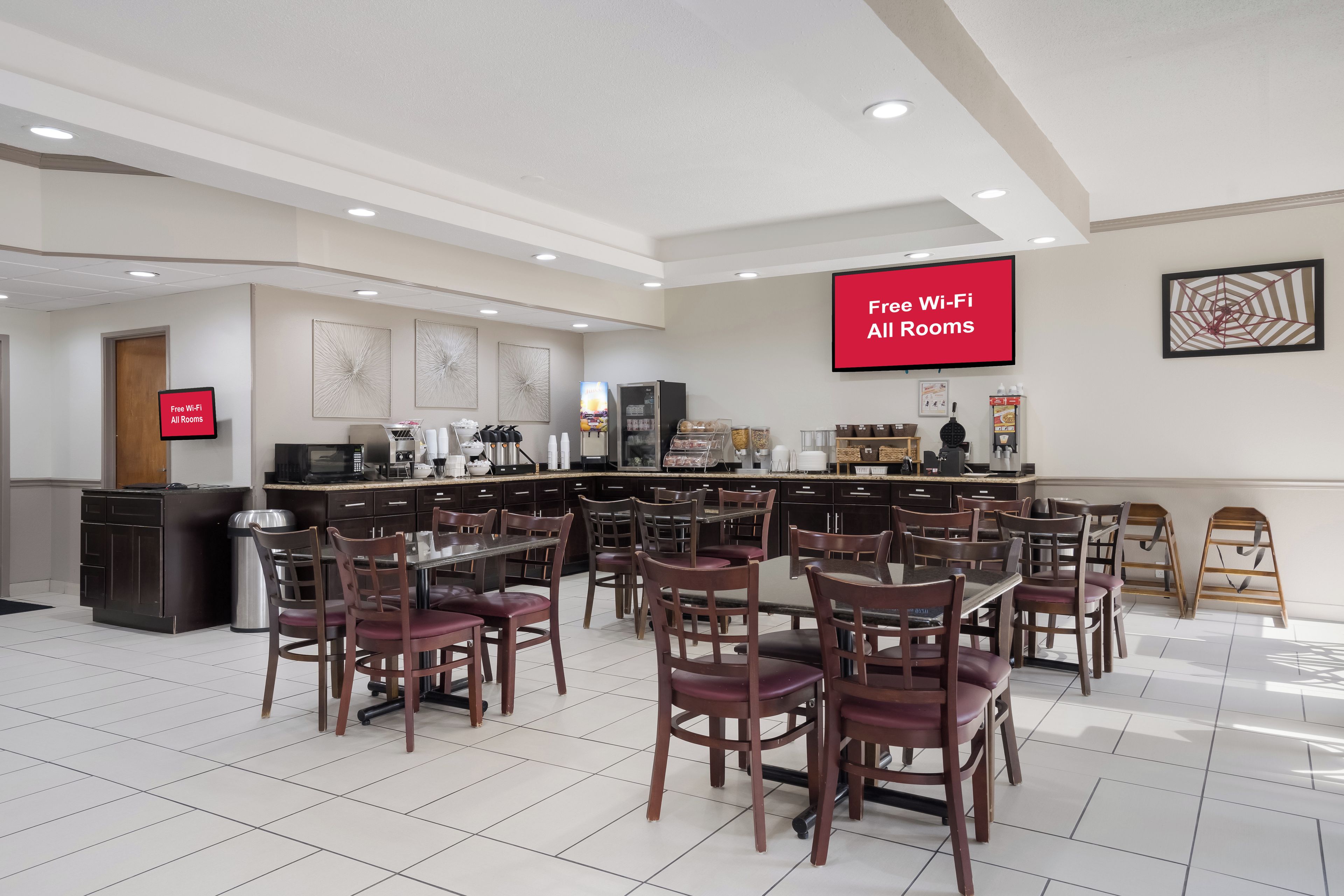 A hotel dining area with multiple tables and chairs, a kitchen area, and a large screen displaying 'Free Wi-Fi All Rooms'.
