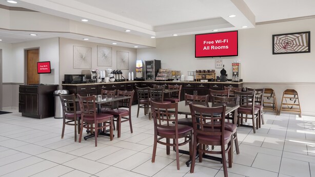 Een restaurantgedeelte in een hotel met meerdere tafels en stoelen, een keuken en een groot scherm waarop 'Gratis Wi-Fi in alle kamers' staat.