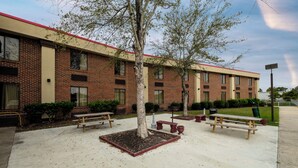 A building with a brick facade, multiple windows, and a red roof. There are outdoor benches and a tree in the courtyard. - Red Roof Inn & Suites Jacksonville, NC (Jacksonville)