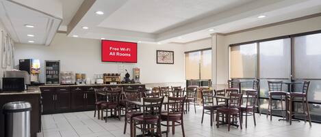 A spacious room with multiple tables and chairs, a kitchen area with a refrigerator and coffee machine, and a large screen displaying 'Free Wi-Fi All Rooms'.