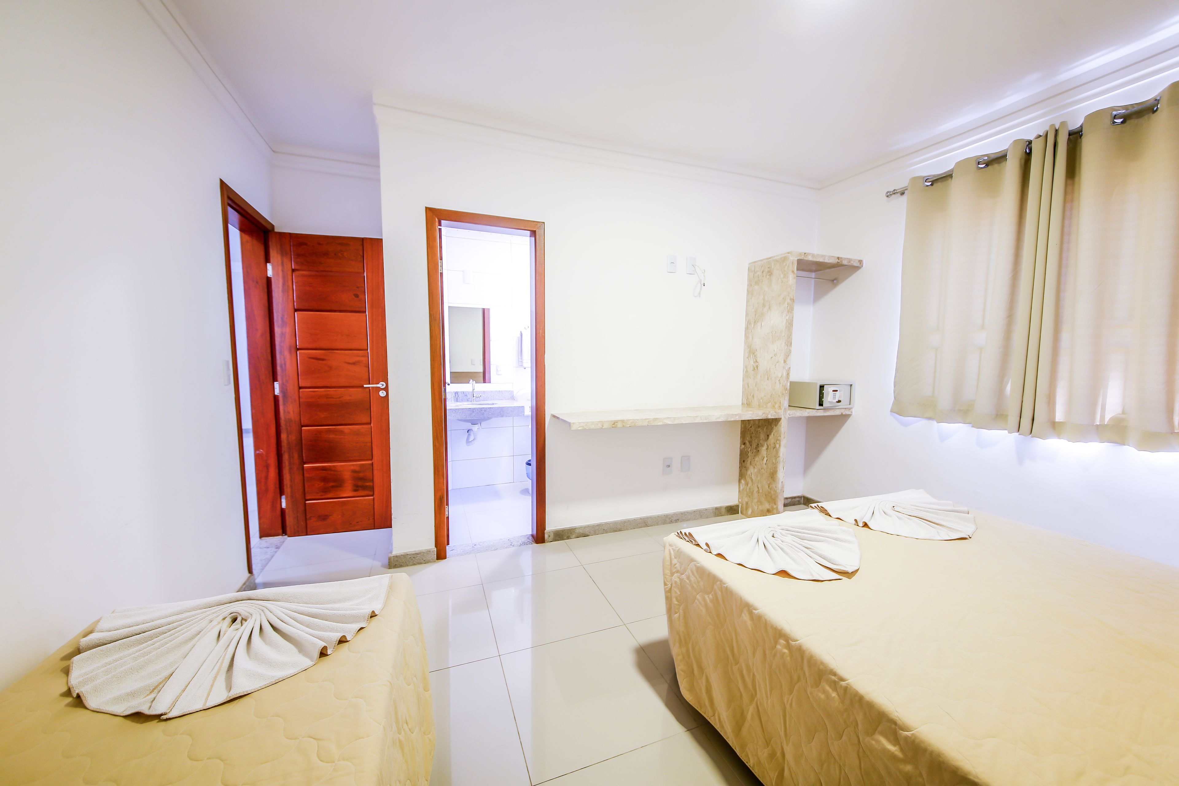 Habitación de hotel con dos camas, una puerta de madera, un baño a la vista a través de una puerta abierta y una ventana con cortinas.