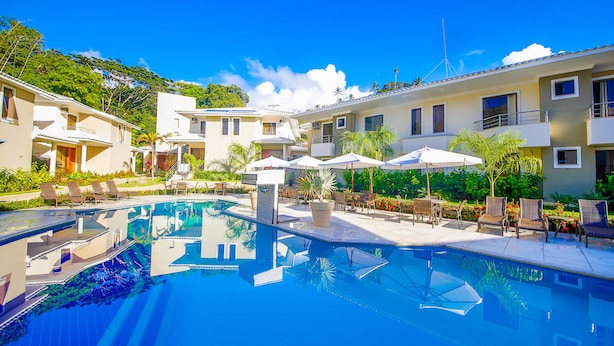 A swimming pool with lounge chairs and umbrellas, surrounded by buildings with balconies and a clear sky.