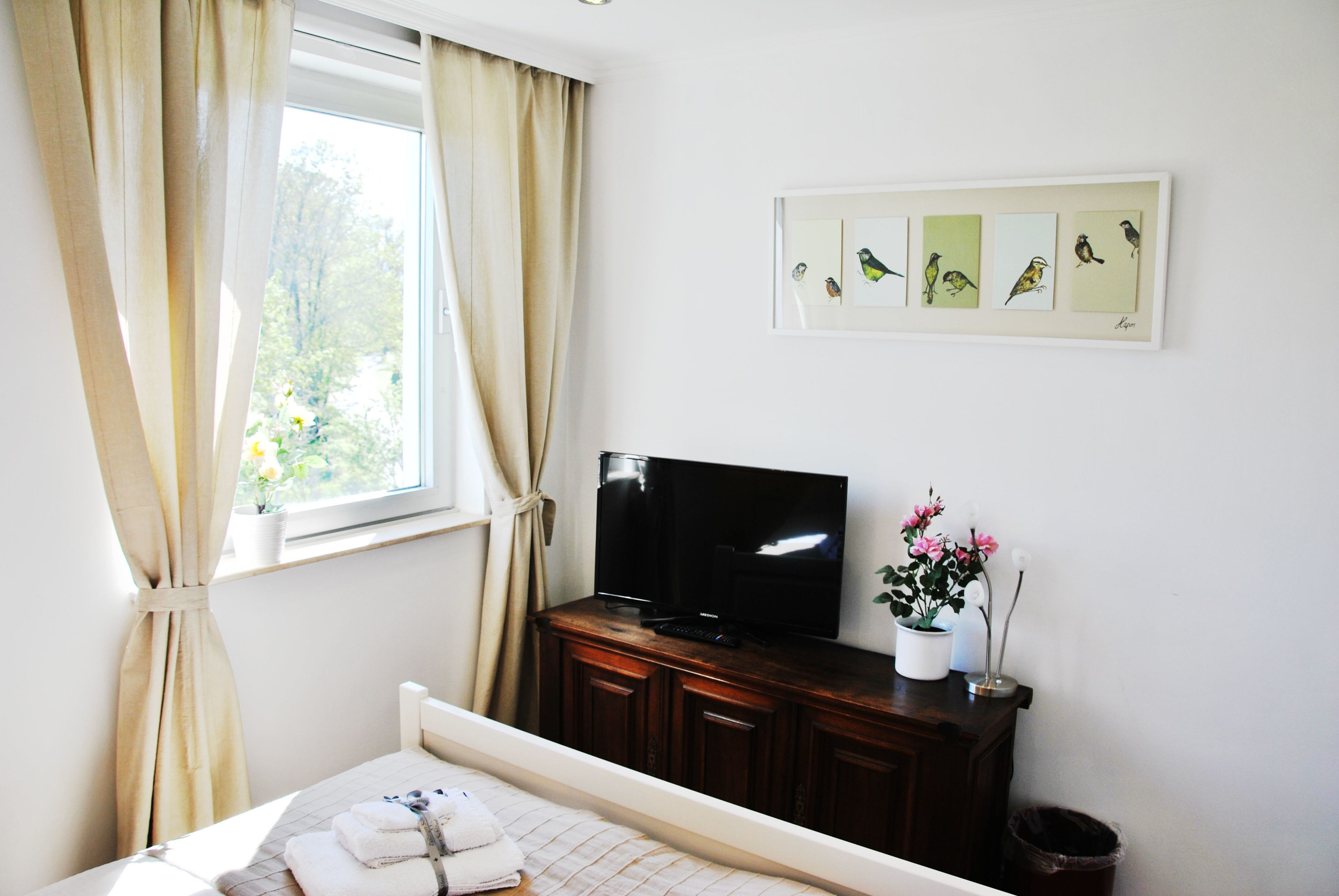 Un dormitorio con cama, televisor sobre un mueble de madera, ventana con cortinas e imagen colgada en la pared de pájaros.