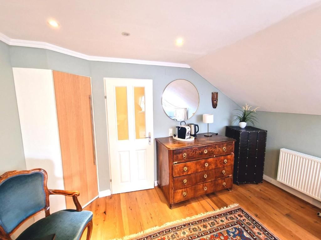 Una habitación con piso de madera, una silla azul, un tocador con cajones, un espejo, un radiador y una puerta.