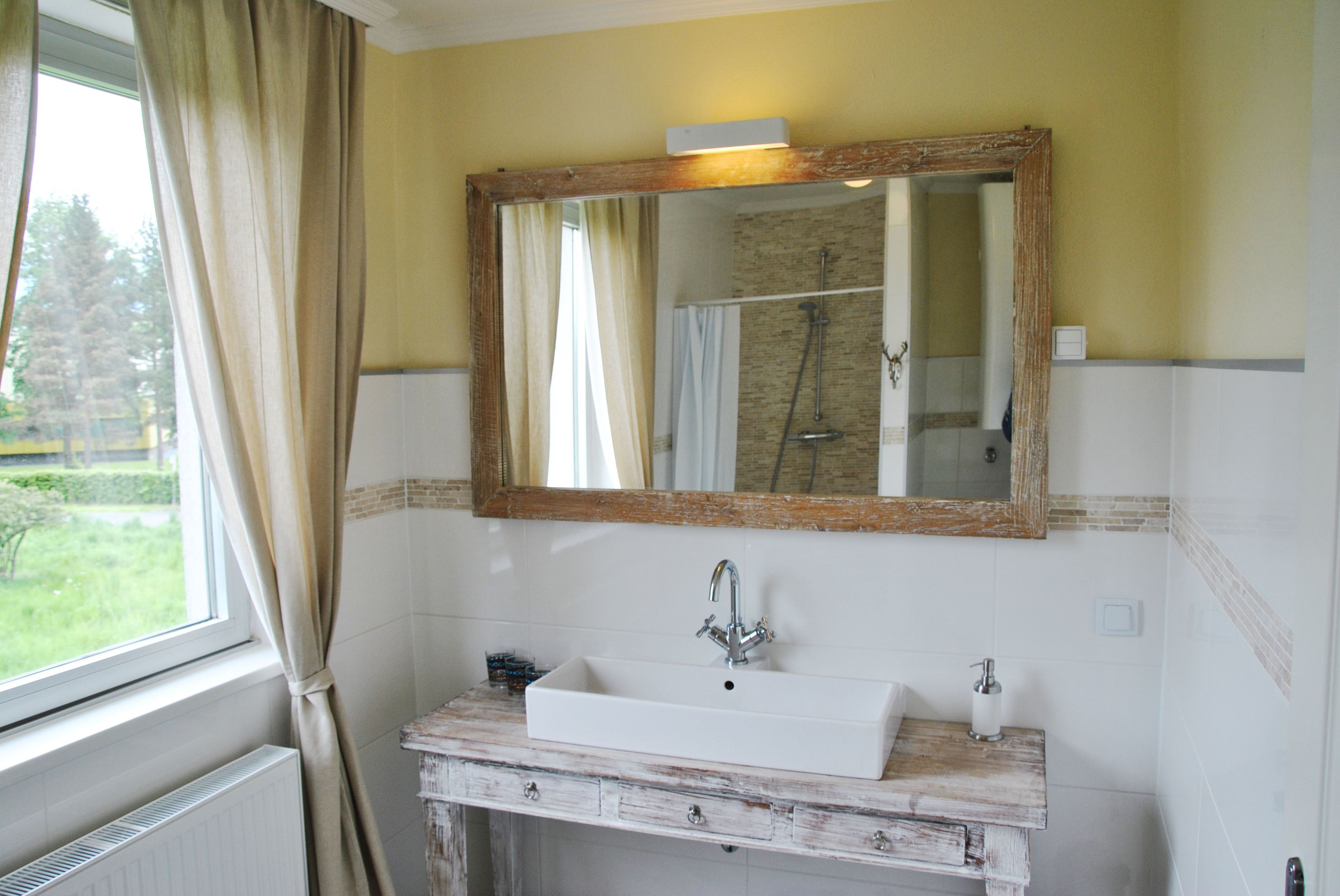 A bathroom with a white sink, a wooden-framed mirror, and a view of a garden through the window.