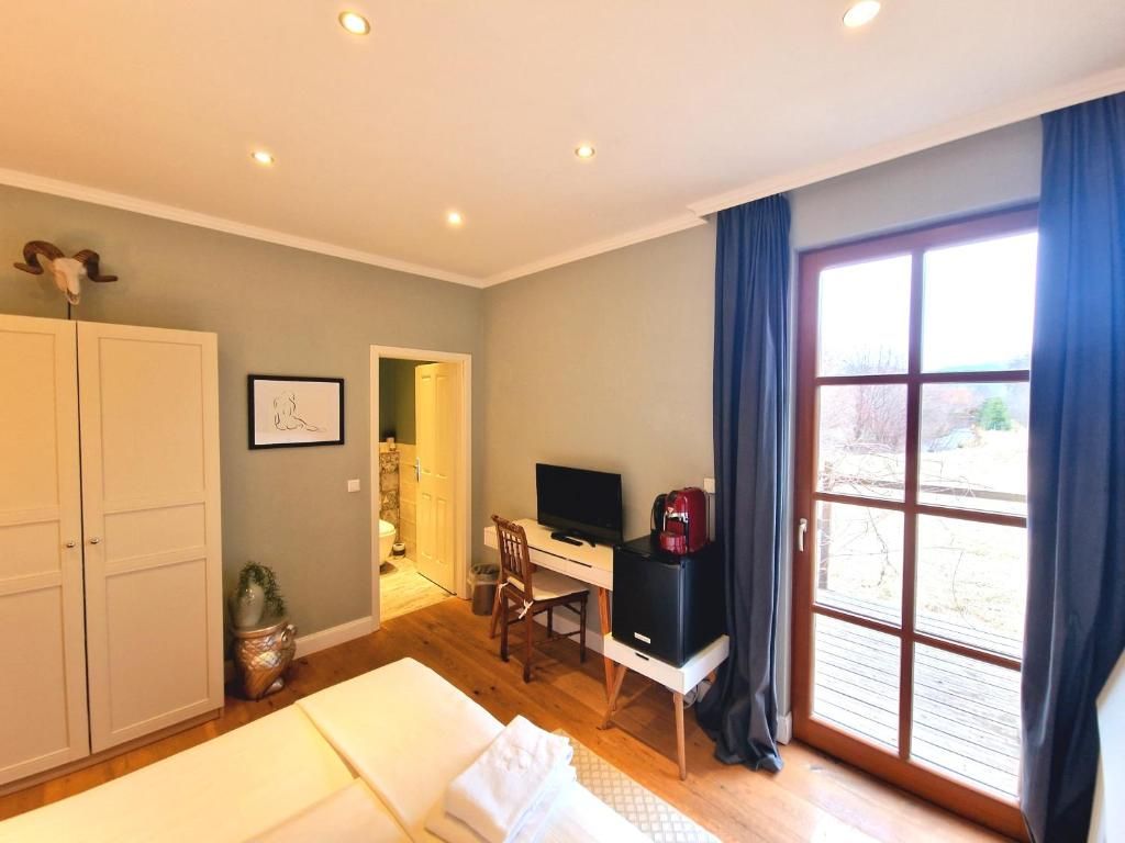 Una habitación con una cama, un escritorio con computadora, un televisor y un amplio ventanal con cortinas.