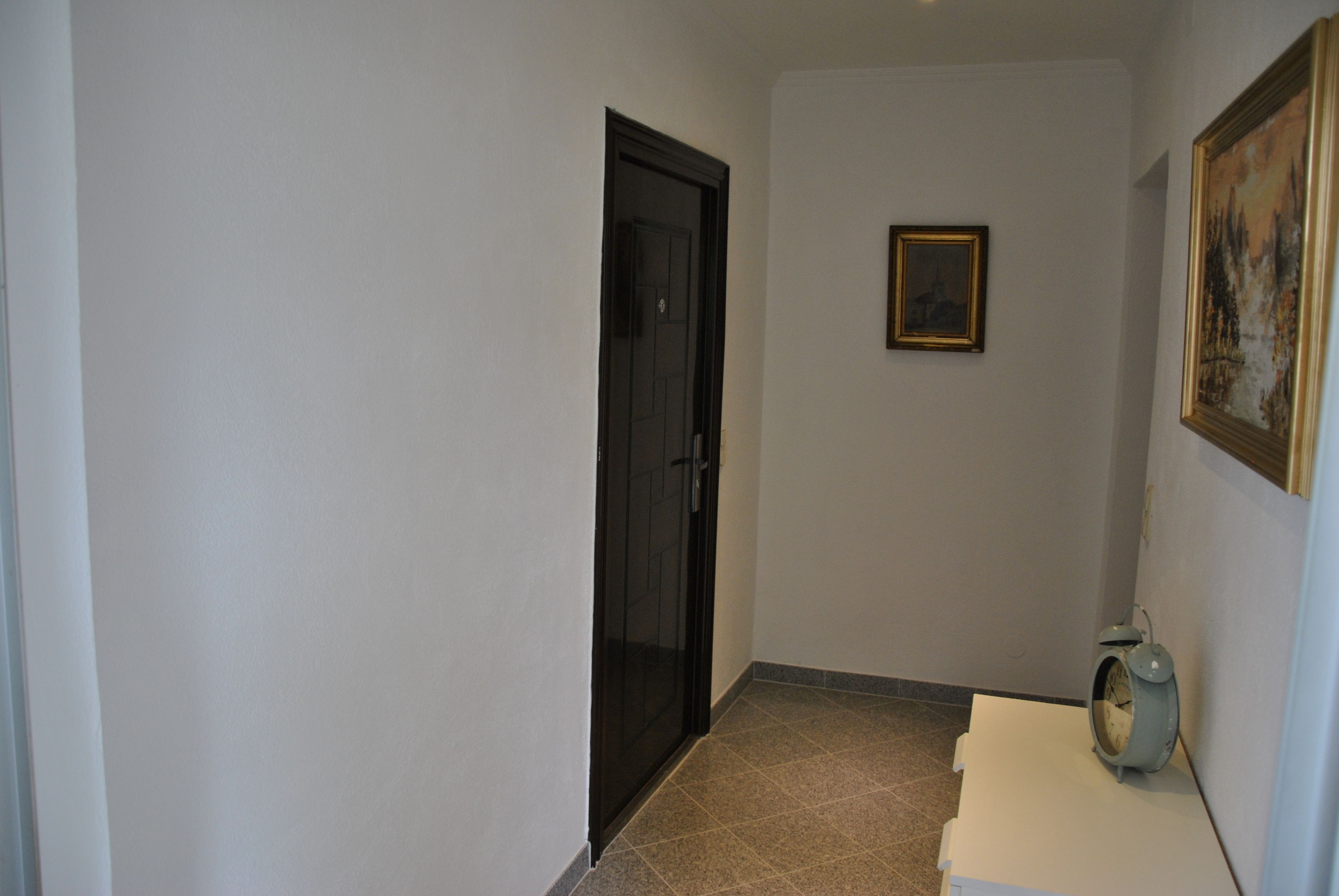 A hallway with a closed door, a framed picture on the wall, and a clock on a table.