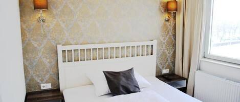 Quarto de hotel com cama branca, cabeceira, duas mesas de cabeceira, um criado-mudo, uma luminária e uma janela com cortinas.