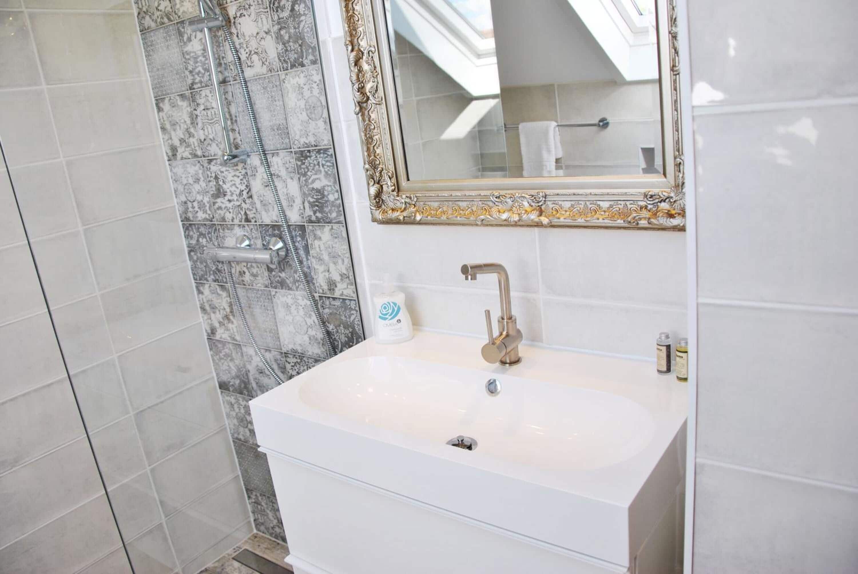 Un baño con un lavabo blanco, un espejo con marco dorado y una zona de ducha con azulejos estampados.